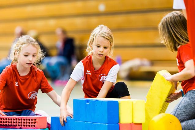 Drei Mädchen im roten Shirt bauen mit bunten Schaumstoffbausteinen in einer Turnhalle.
