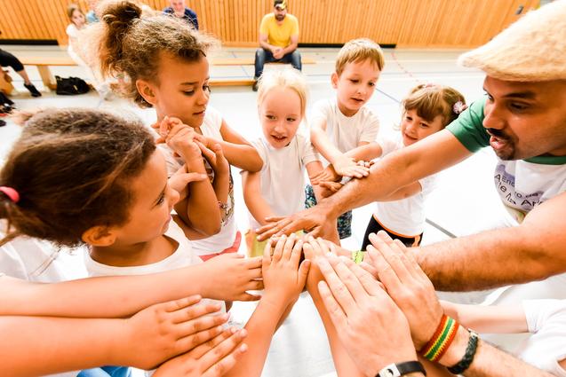 Gruppenspiel mit Kindern, die ihre Hände zum gemeinsamen Aufeinandertreffen heben, unter Anleitung eines Erwachsenen.