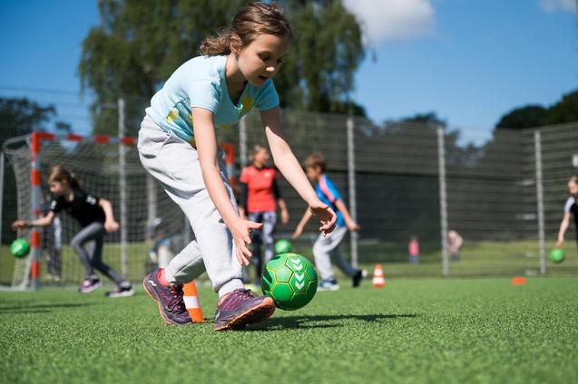 Mädchen in Sportkleidung spielt mit einem grünen Ball auf einem Kunstrasenplatz, andere Kinder trainieren im Hintergrund.